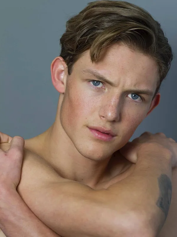 Portrait d'un jeune homme torse nu aux yeux bleus, regardant l'objectif, les bras croisés derrière la tête.