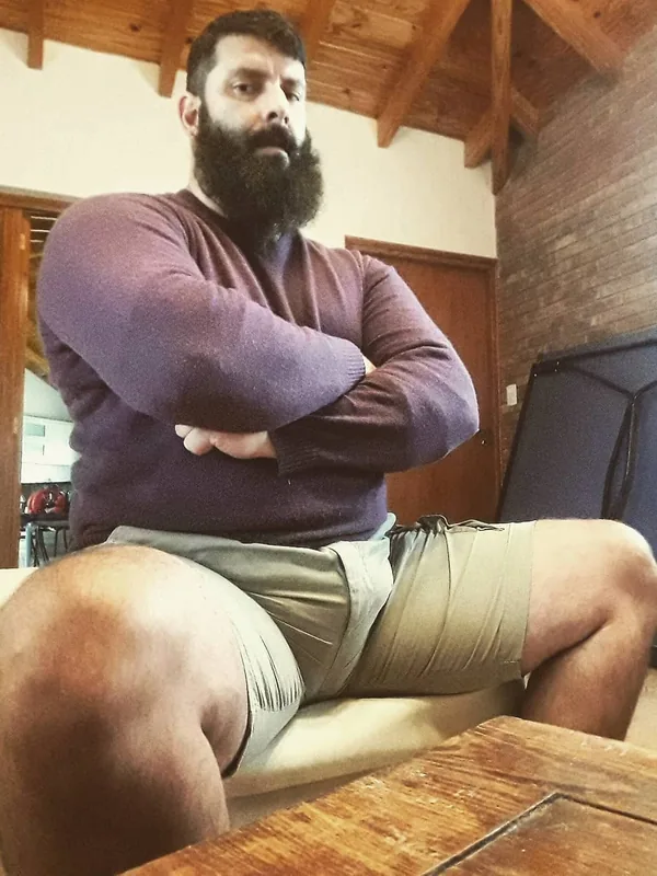 Homme barbu en pull violet et shorts gris assis sur un canapé dans une pièce avec poutres en bois.