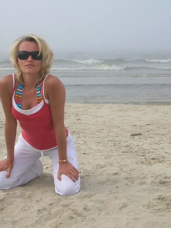 Femme blonde en tenue claire agenouillée sur une plage, portant des lunettes de soleil, devant l'océan sous un ciel brumeux.