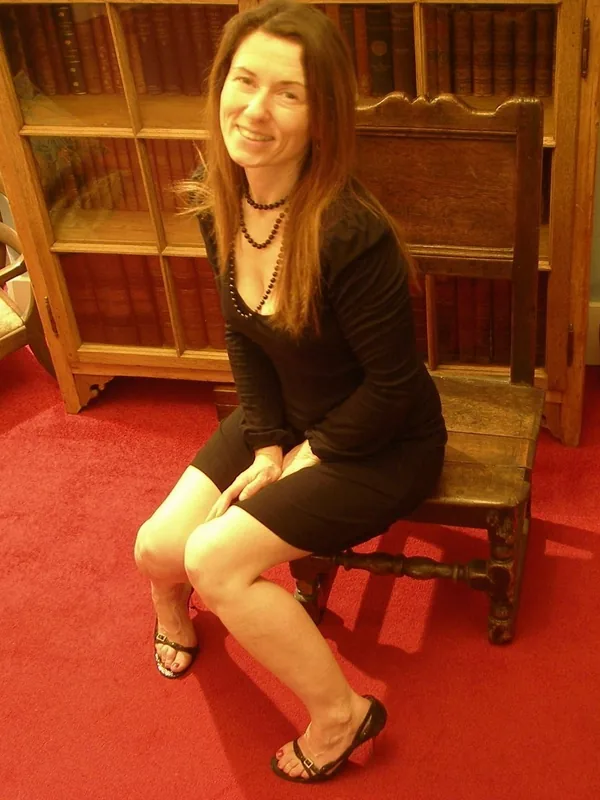Femme souriante assise sur une chaise en bois devant une bibliothèque, vêtue d'une robe noire et de sandales à talons.