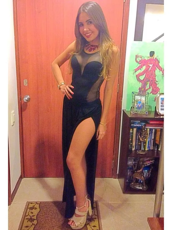 Femme souriante en robe noire à fente haute, posant en pied devant une porte en bois dans un intérieur.