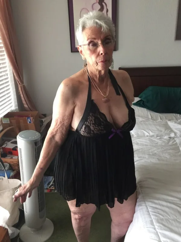 Femme âgée aux cheveux blancs portant une robe noire à dentelle, debout dans une chambre près d'un lit et d'un ventilateur.