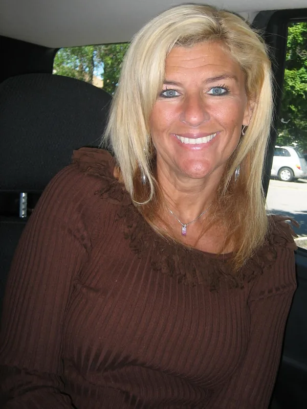 Femme blonde souriante assise à l'arrière d'une voiture, portant un haut marron et un pendentif rose.