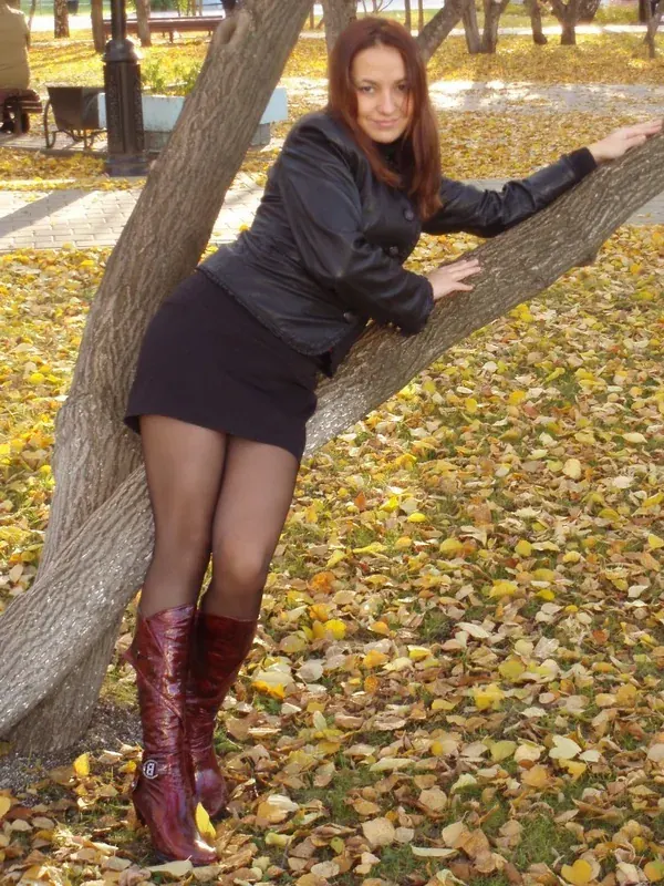 Femme en veste en cuir et bottes rouges appuyée contre un arbre parmi des feuilles d'automne.