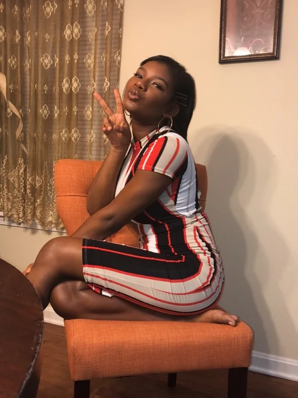 Femme assise sur une chaise orange, portant une robe rayée, faisant un signe de la main, dans un intérieur éclairé.