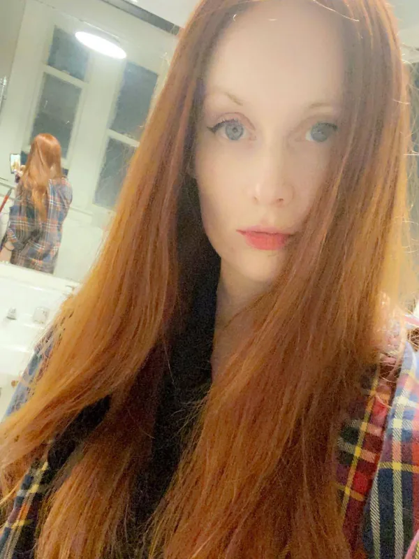 Portrait en gros plan d'une femme aux cheveux roux portant une chemise à carreaux, prise devant un miroir dans une pièce éclairée.