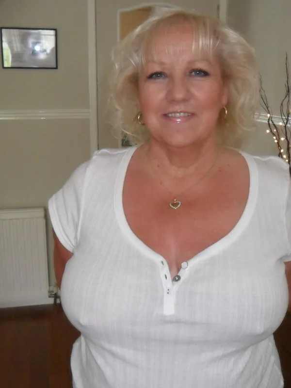 Portrait en buste d'une femme blonde souriante portant un haut blanc, dans un intérieur éclairé.