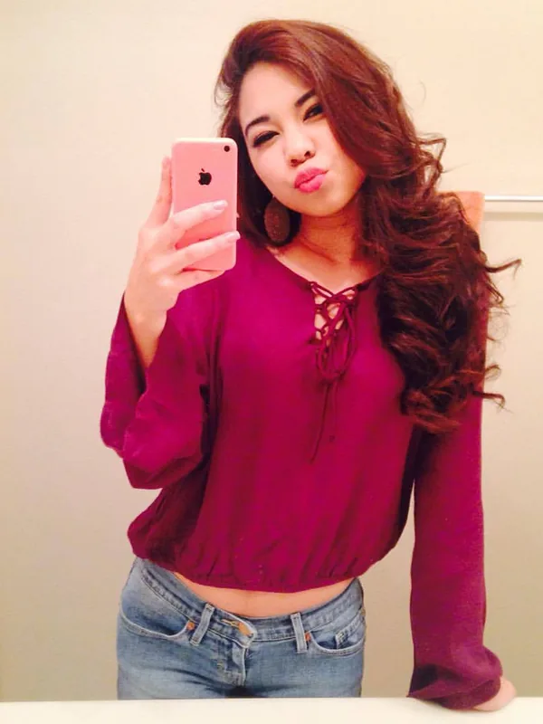 Femme aux cheveux bruns ondulés prenant un selfie avec un téléphone rose devant un miroir, portant une chemise violette et un jean.