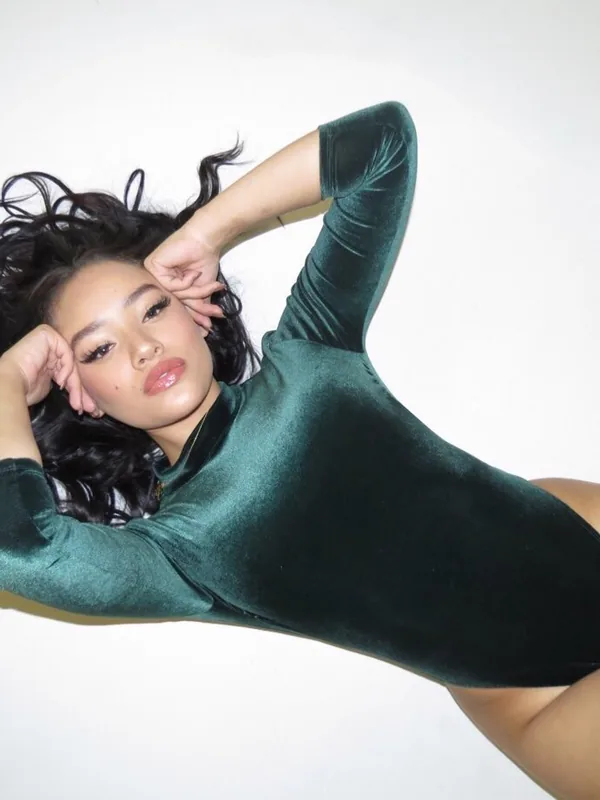 Femme allongée sur le côté, portant un body vert foncé en velours, les mains derrière la tête, sur fond blanc.