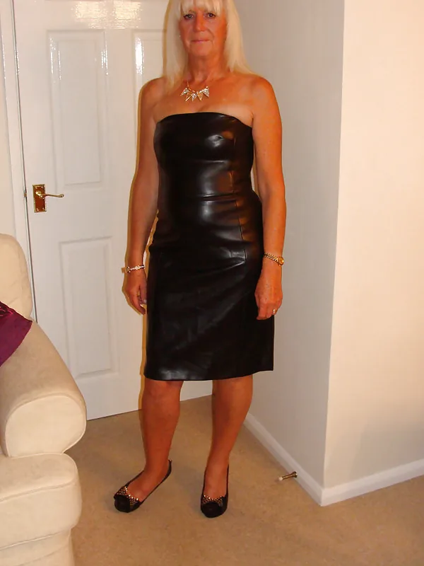Femme blonde en robe noire sans bretelles en cuir, debout dans un salon, regardant l'objectif.