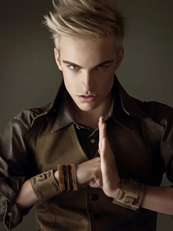 Portrait d'un jeune homme aux cheveux blonds portant une veste marron et des bracelets, les mains jointes, regardant l'objectif.