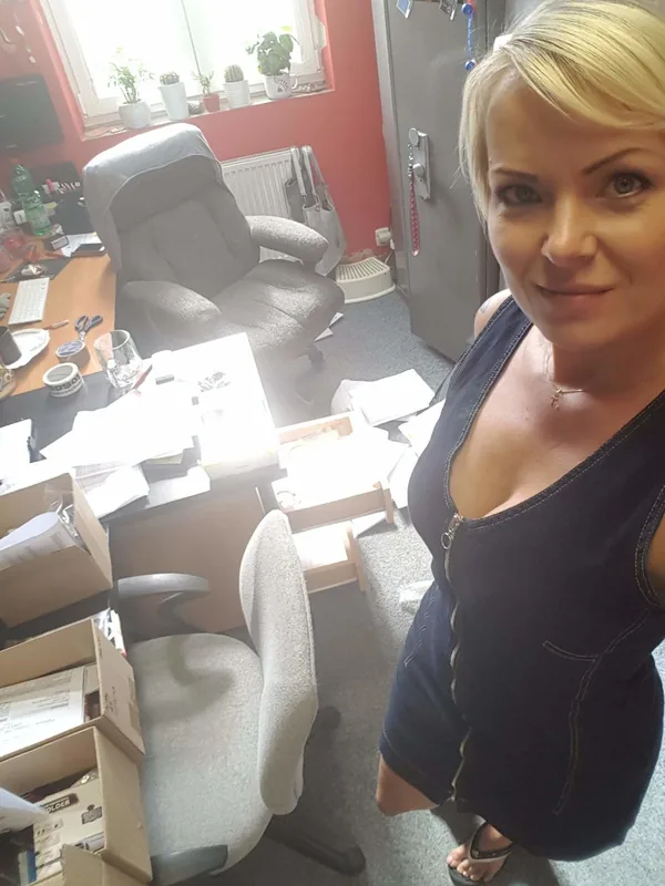 Femme blonde en robe noire posant dans un bureau en désordre avec des boîtes et des papiers.