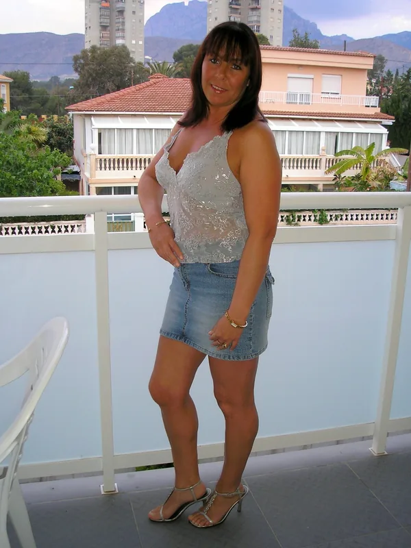 Femme debout sur un balcon, portant un haut transparent et un short en jean, regardant l'objectif.