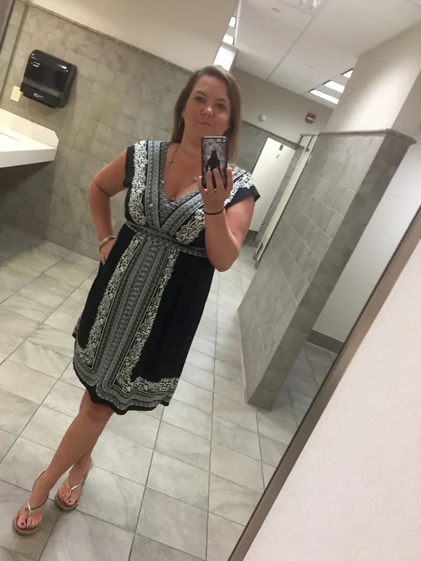 Femme prenant un selfie dans un miroir d'une salle de bain, vêtue d'une robe noire à motifs et de sandales.