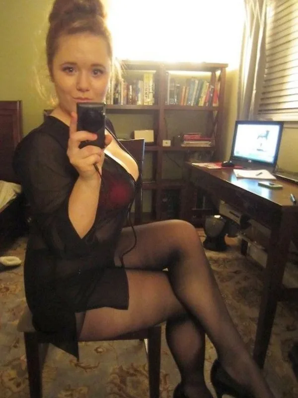 Femme assise sur une chaise, prenant un selfie avec un téléphone, vêtue d'une robe noire transparente et de collants, dans une chambre.