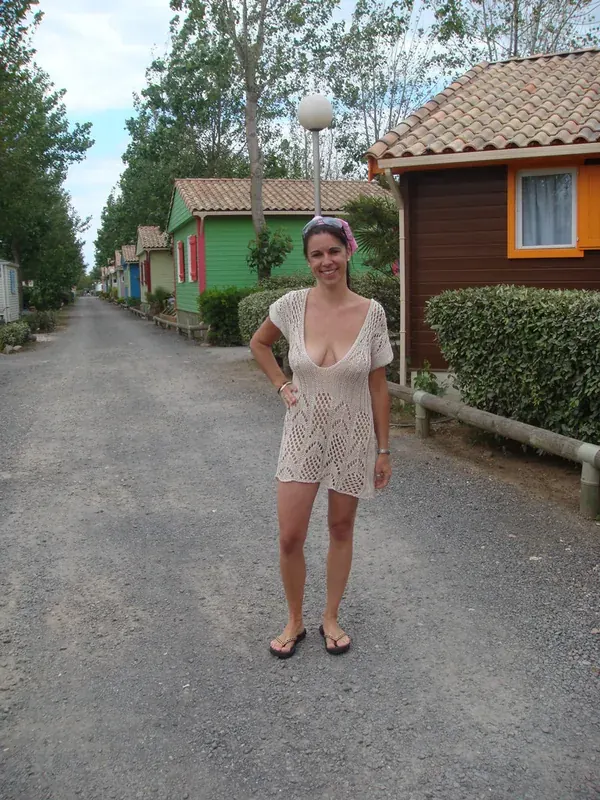 Femme souriante en robe en crochet beige, debout sur une allée entre des chalets colorés dans un camping.