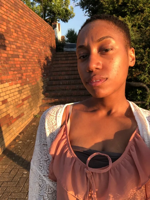 Portrait d'une femme aux cheveux courts, en haut rose et veste blanche, devant un mur de briques au coucher du soleil.