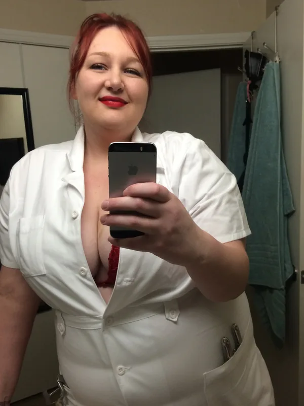 Femme aux cheveux roux prenant un selfie dans un miroir, vêtue d'une blouse blanche ouverte sur un soutien-gorge rouge.