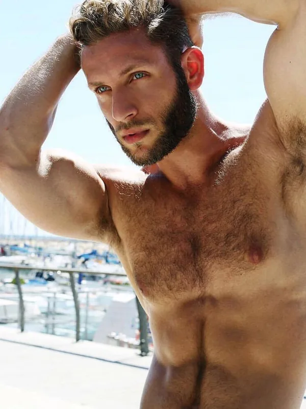 Portrait d'un homme torse nu aux cheveux bruns et barbe, regardant l'objectif, devant un port ensoleillé.