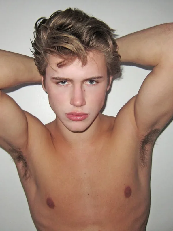 Portrait d'un jeune homme torse nu, cheveux blonds, bras derrière la tête, regardant l'objectif sur fond uni.