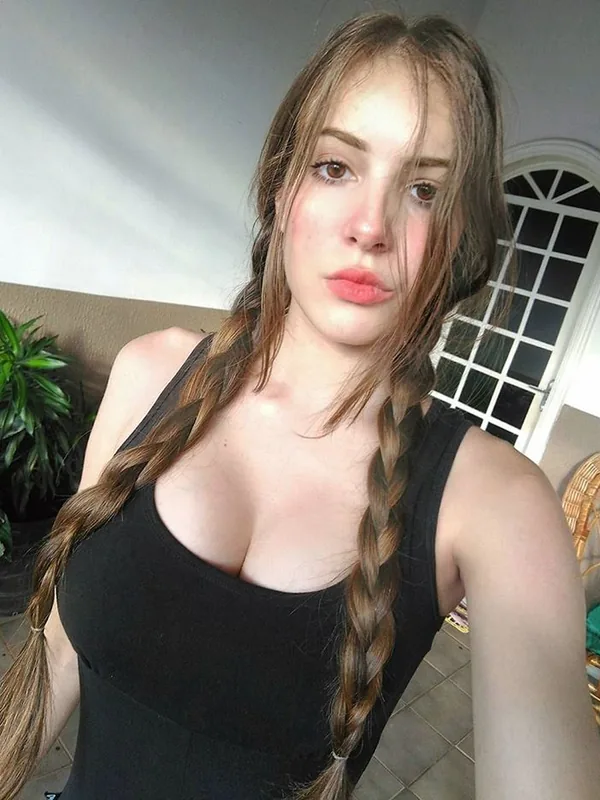 Portrait d'une jeune femme aux cheveux bruns en tresses, portant un haut noir, regardant l'objectif dans un intérieur lumineux.