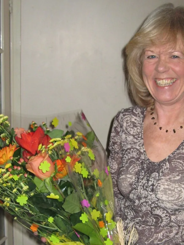 Femme souriante tenant un grand bouquet de fleurs colorées devant elle, en intérieur.