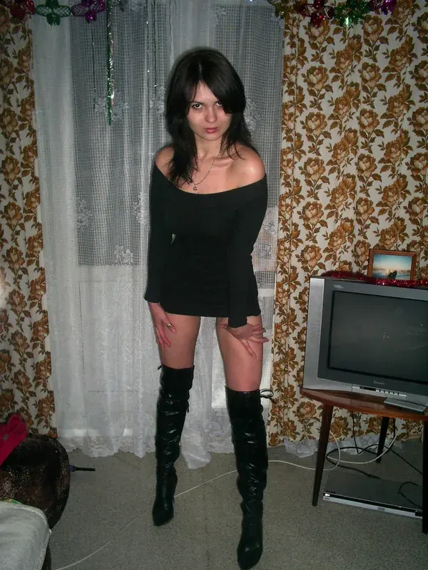 Femme aux cheveux noirs en tenue noire et bottes hautes, debout devant rideaux et papier peint floral.