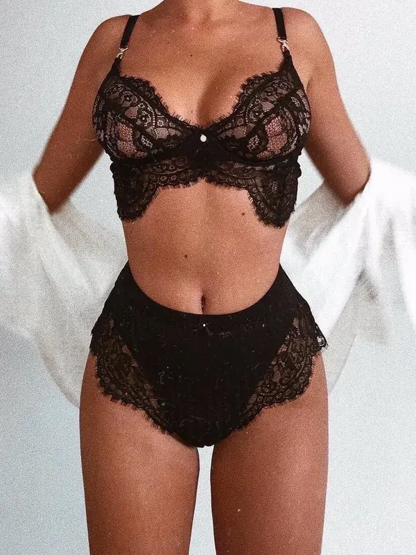 Femme portant un ensemble de lingerie noire en dentelle, avec un vêtement blanc ouvert sur les épaules, devant un fond clair.