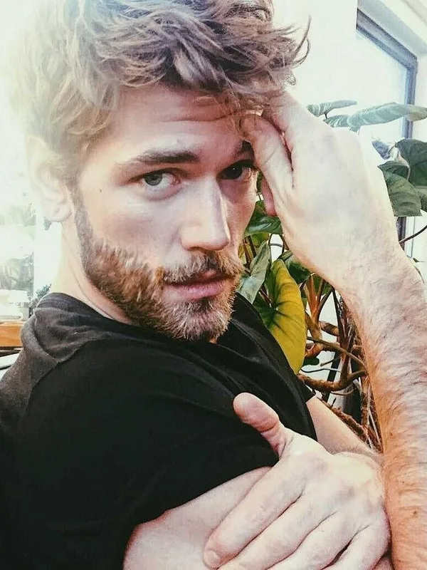 Portrait d'un homme aux cheveux bruns et barbe, portant un t-shirt noir, regardant l'objectif, avec une plante en arrière-plan.