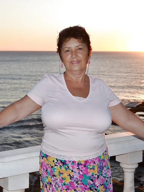 Femme souriante en tenue claire et jupe fleurie, appuyée sur une balustrade face à la mer au coucher du soleil.