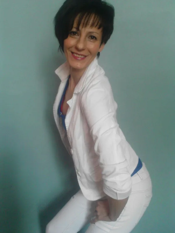 Femme aux cheveux courts noirs, souriante, vêtue d'une chemise blanche et de jeans blancs, posant contre un mur turquoise.
