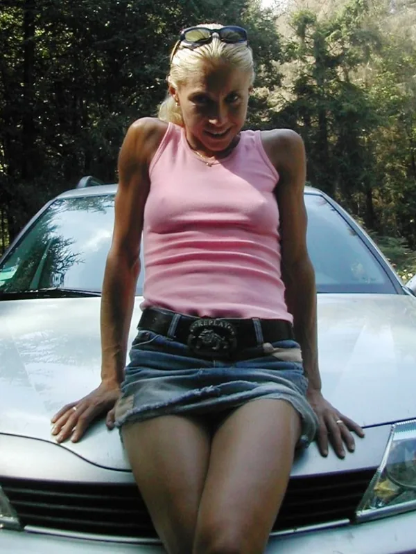 Femme blonde en haut rose et jupe en jean assise sur le capot d'une voiture blanche en extérieur.