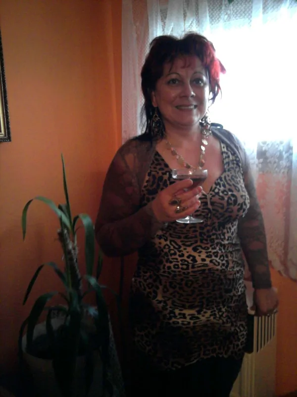 Femme souriante tenant un verre de vin rouge, vêtue d'un haut léopard et d'une veste transparente, devant une fenêtre avec rideaux blancs.