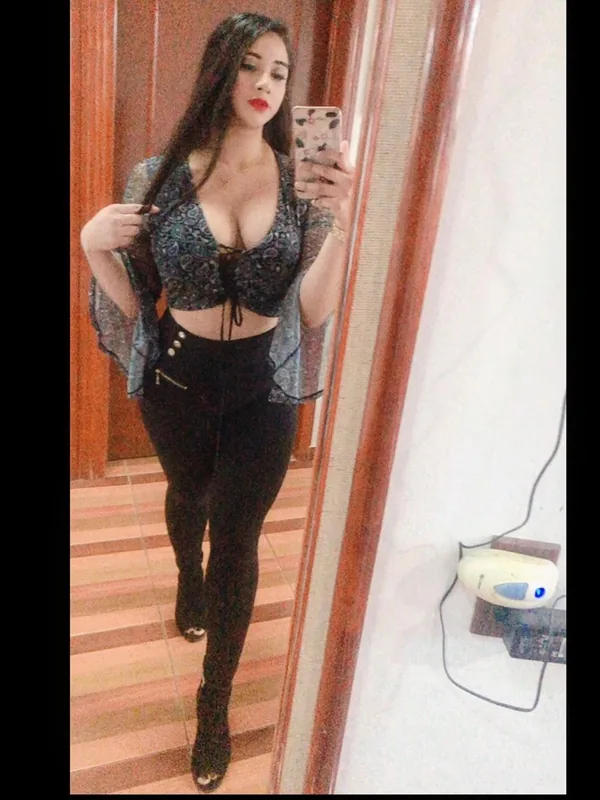 Portrait de femme prenant un selfie devant un miroir, vêtue d'un haut à motifs et de leggings noirs, dans un couloir intérieur.