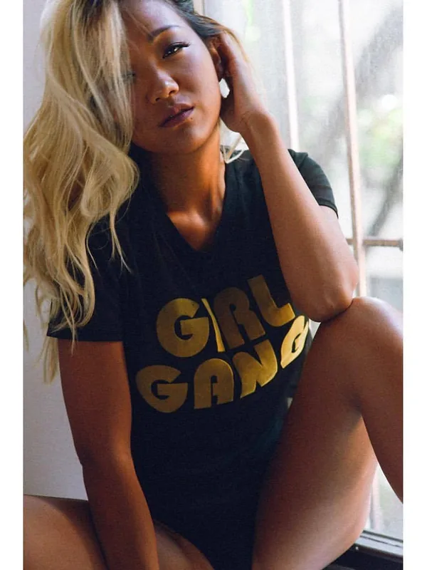 Femme aux cheveux blonds portant un t-shirt noir avec inscription 'GIRL GANG', assise près d'une fenêtre, regardant vers l'objectif.