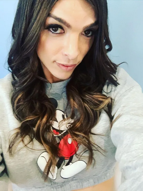 Portrait d'une femme aux cheveux bruns ondulés portant un haut gris à motif Mickey Mouse, regardant l'objectif.