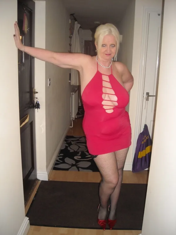 Femme blonde en robe rose avec découpes, collants noirs et chaussures rouges, posant dans un couloir intérieur.