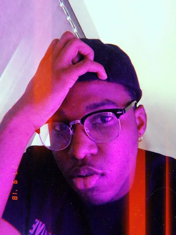 Portrait d'un homme portant une casquette et des lunettes, la main sur le front, sous une lumière violette et rose.