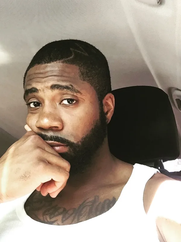 Homme à la barbe et au tatouage sur le torse, portant un débardeur blanc, assis dans une voiture et regardant l'objectif.